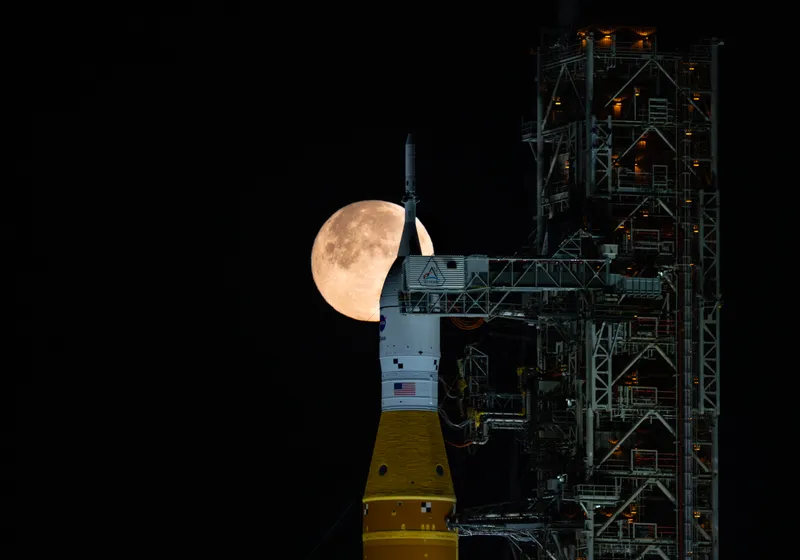 A full moon rising beside the Artemis II rocket on Launch Pad 39B