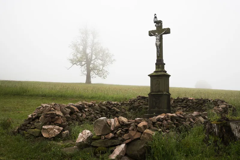 A stone wayside cross in morning mist with a tree behind it